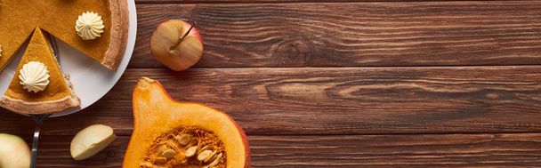 panoramic shot of delicious pumpkin pie with whipped cream near cut and whole apples, half of raw pumpkin on brown wooden table - Photo, Image
