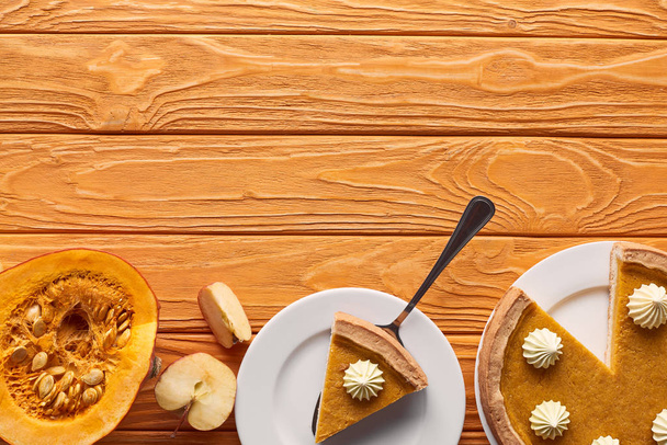 おいしいカボチャのパイカットの近くにホイップクリームとリンゴ全体,オレンジの木のテーブルの上に生のカボチャの半分 - 写真・画像