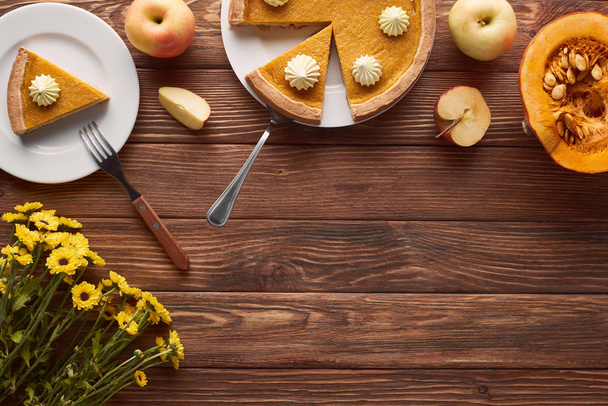 tasty pumpkin pie with whipped cream near spatula and fork, half of raw pumpkin, whole and cut apples on brown wooden surface - Foto, afbeelding