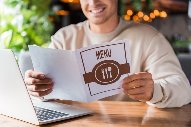 ostříhaný pohled na šťastného muže držícího menu v blízkosti notebooku v restauraci  - Fotografie, Obrázek