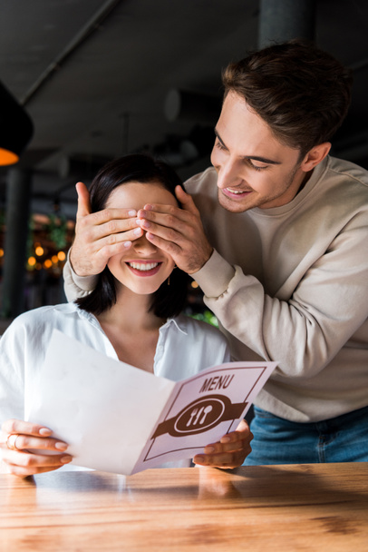 happy man closing eyes of cheerful woman holding menu in restaurant  - Foto, Imagen