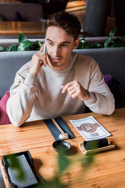selective focus of handsome man talking on smartphone in sushi bar near table with menu  - Fotó, kép