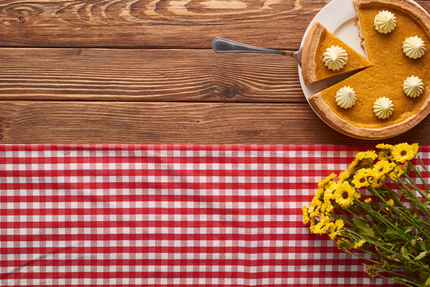вырезанный тыквенный пирог со взбитыми сливками возле букета желтых цветов и клетчатой скатерти на деревянном столе
 - Фото, изображение