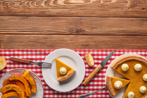 cut pumpkin pie with whipped near baked sliced pumpkin, fork and knife of checkered tablecloth on wooden surface - Photo, Image