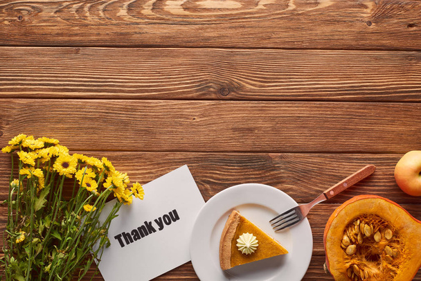 piece of pumpkin pie with whipped cream near thank you greeting card, fork, raw pumpkin and yellow flowers on wooden table - Photo, Image