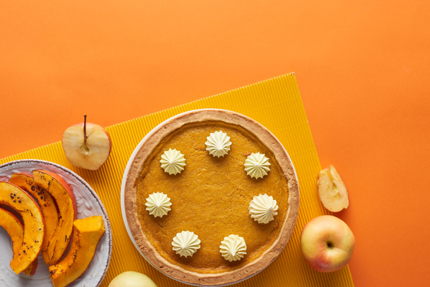 tasty pumkin pie with whipped cream near baked pumpkin, whole and cut apples on orange surface - Photo, image