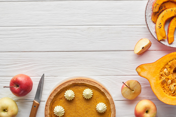 tasty pumpkin pie with whipped cream near baked pumpkin, whole and cut apples, and knife on white wooden surface - 写真・画像