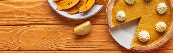 panoramic shot of delicious pumpkin pie with whipped cream near baked pumpkin and cut apple on orange wooden table - 写真・画像