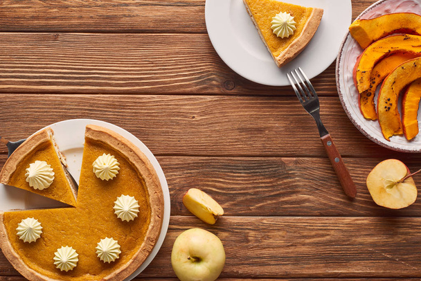 cut pumpkin pie with whipped cream near baked pumpkin, whole and cut apples and fork on brown wooden table - Foto, Bild