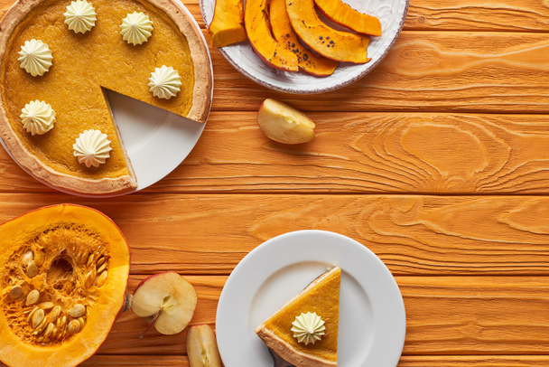 delicious, cut pumpkin pie with whipped cream near baked and raw pumpkins, and cut apple on orange wooden table - Valokuva, kuva