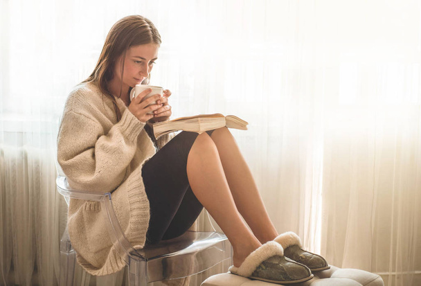 Το όμορφο κορίτσι διαβάζει ένα βιβλίο με ένα φλιτζάνι ζεστό τσάι. Χειμερινή ώρα φθινοπώρου.  - Φωτογραφία, εικόνα