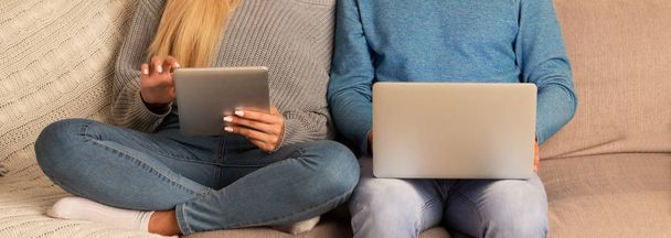 Casal irreconhecível usando computadores portáteis e tablets sentados no sofá
 - Foto, Imagem