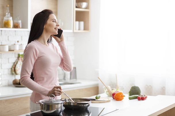 Tysiąclecia kobieta rozmawia na telefon, przygotowuje obiad w kuchni - Zdjęcie, obraz