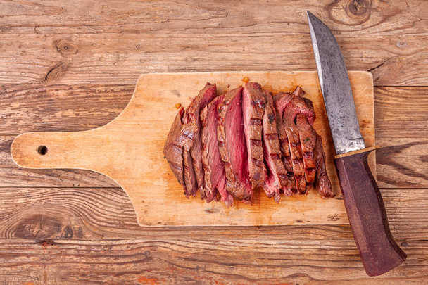 Шматочки смаженого яловичого м'яса на дерев'яній обробній дошці зі старим ножем. Вид зверху
. - Фото, зображення