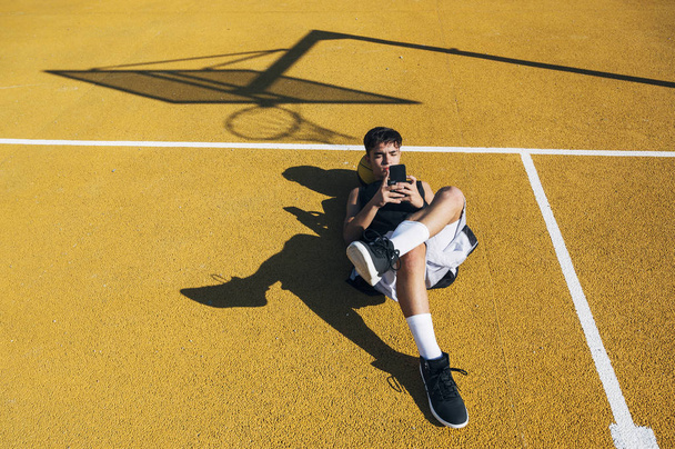 トレーニングセッション後に黄色いコートに横たわっている間、スマートフォンを使用して男性バスケットボール選手. - 写真・画像