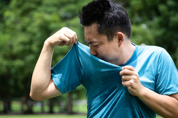 Asijský mladý muž se cítí špatně, něco smrdí, čichá k mokrému podpaží nebo se hodně potí kvůli horkému počasí nebo po cvičení ve venkovním parku, mužský výraz obličeje, koncept použití deodorantu  - Fotografie, Obrázek