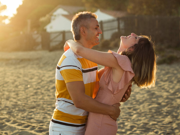 Podekscytowany dorosły mężczyzna i kobieta śmieją się i przytulają podczas zabawy na piaszczystym brzegu w kurorcie - Zdjęcie, obraz