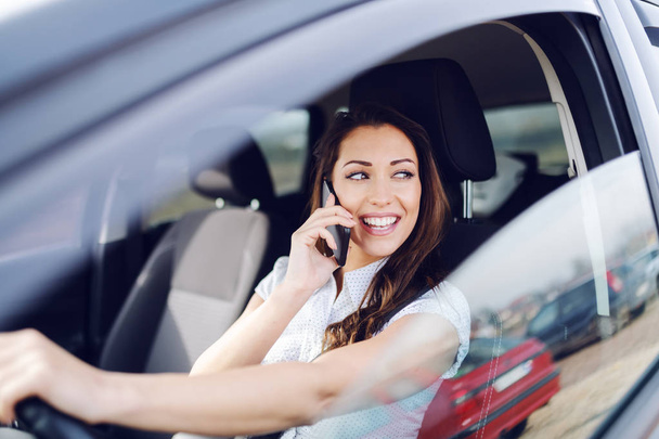 Houkutteleva valkoihoinen hymyilevä brunette ajaa autonsa ja puhuu puhelimessa katsellen kaukalo ikkuna
. - Valokuva, kuva