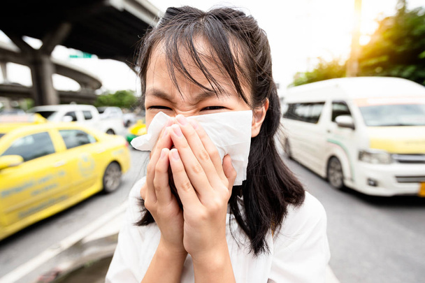 Portrait-Nahaufnahme eines Kindes, das Nase in Papiertaschentuch pustet, asiatische Frau, die in ein Taschentuch in der Stadtstraße niest, giftige Dämpfe aus dem Auto, schlechter Geruch, Begriff der Luftverschmutzung, Stauballergien, Gesundheitsfürsorge  - Foto, Bild