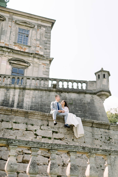 Beautiful Asian wedding couple poses while sitting on the stone wall of an old ruined castle - Foto, Imagen