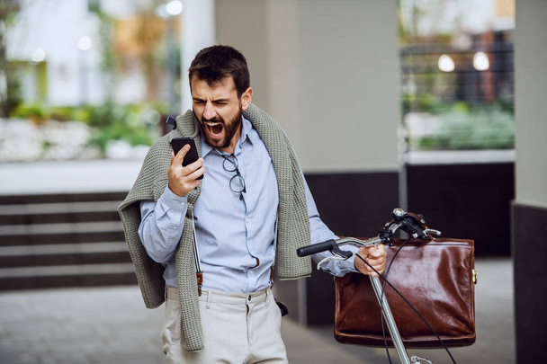 Nervous bearded fashionable caucasian employee pushing his bike, holding smart phone and yelling. Overtime concept. - Фото, зображення