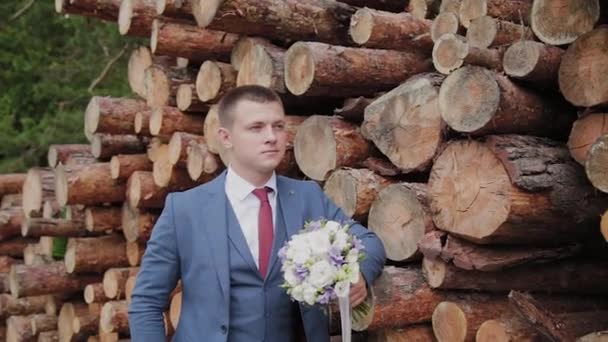 schöner Bräutigam in Jacke und mit einem Strauß steht neben einem Stapel Baumstämme. - Filmmaterial, Video