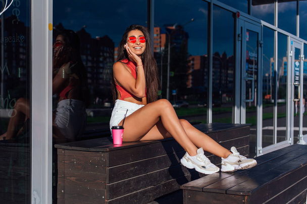 Stylish happy young brunette woman wearing white shorts and sneakers holding pink cup of coffee to go sitting next to coffee shop. - Photo, Image