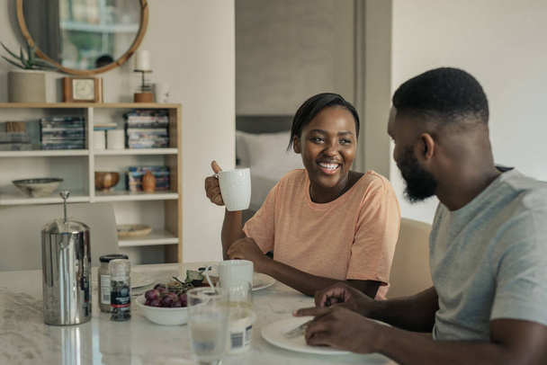 Young African American woman laughing while drinking coffee and eating breakfast with her husband at home in the morning - 写真・画像