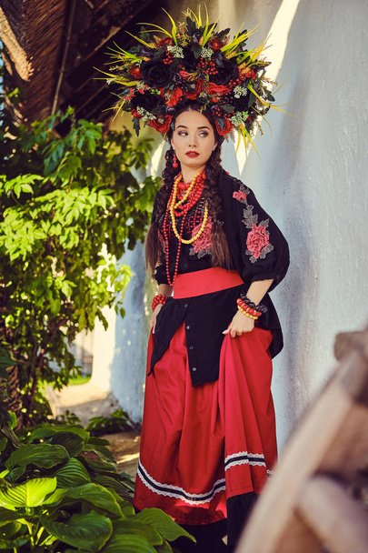 Брюнетка дівчина в чорному і червоному вишитому українському автентичному національному костюмі і вінок з квітів позує стоячи проти білої хатини. - Фото, зображення