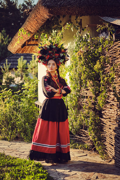Μελαχρινή κορίτσι σε μαύρο και κόκκινο κεντημένο ουκρανικό αυθεντικό εθνικό κοστούμι και ένα στεφάνι από λουλούδια ποζάρει στέκεται εναντίον μιας λευκής καλύβας. - Φωτογραφία, εικόνα