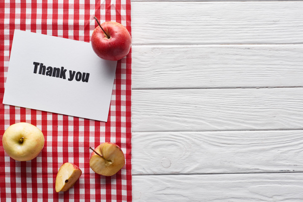 Draufsicht auf reife Äpfel und Dankeskarte auf weißem Holztisch mit rot karierter Serviette - Foto, Bild