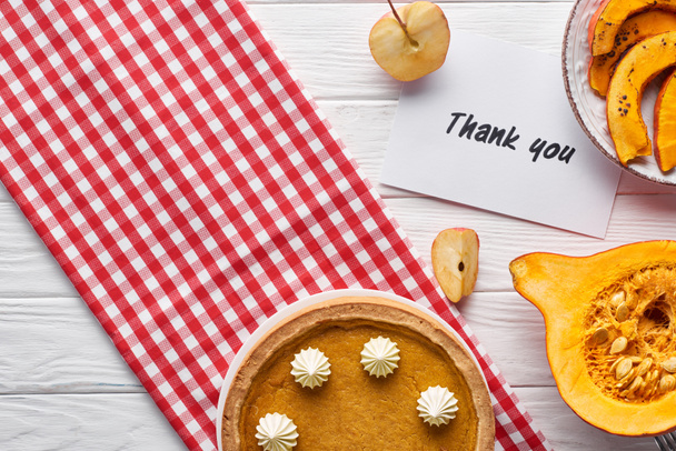 Ansicht von Kürbiskuchen, reifen Äpfeln und Dankeskarte auf weißem Holztisch mit karierter Serviette - Foto, Bild