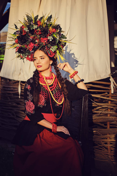 Μελαχρινή κορίτσι σε ένα μαύρο και κόκκινο κεντημένο ουκρανικό αυθεντικό εθνικό κοστούμι και ένα στεφάνι από λουλούδια ποζάρει σε μια βεράντα. Γκρο πλαν. - Φωτογραφία, εικόνα