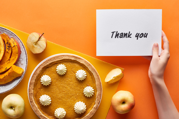 oříznutý pohled na ženu s poděkováním kartičky v blízkosti dýňového koláče, zralých jablek na oranžovém pozadí - Fotografie, Obrázek