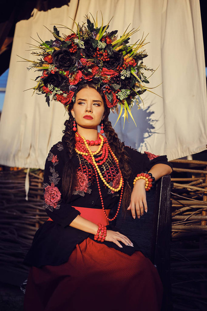Дівчина брюнетка в чорно-червоному вишитому українському автентичному національному костюмі і вінок з квітів позує на терасі. Закри. - Фото, зображення