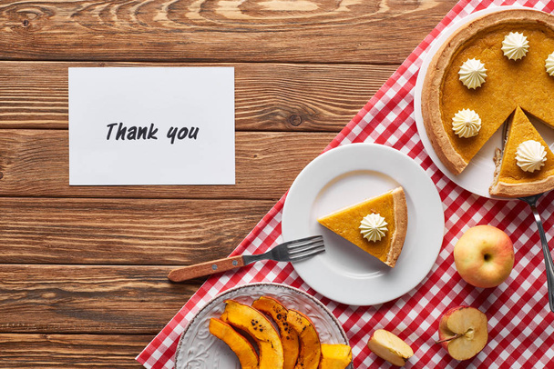 top view of pumpkin pie, ripe apples and thank you card on wooden brown table with red plaid napkin - Photo, Image