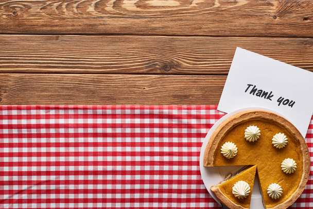 Draufsicht auf Kürbiskuchen und Dankeskarte auf hölzernem braunem Tisch mit rot karierter Serviette - Foto, Bild