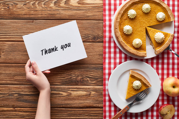 cropped view of woman holding thank you card near pumpkin pie, ripe apples on plaid napkin on wooden table - Photo, Image