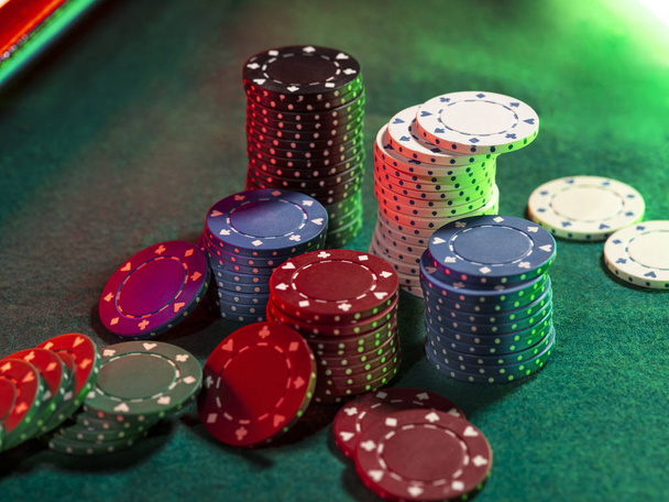 Close-up widok z góry kolorowe stosy żetonów, niektóre z nich leżące w pobliżu na zielonej okładce stołu do gry, pod zielonym, czerwonym świetle neonu. Czarne tło. - Zdjęcie, obraz