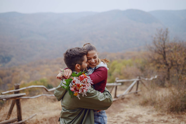 Feliz pareja multicultural abrazándose en la naturaleza. Hombre sosteniendo a su novia en brazos y mujer sosteniendo ramo. Temporada de otoño
. - Foto, Imagen