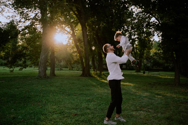 Bij zonsondergang vader met zijn kleine jongen spelen in het park het nemen van zijn zoon in handen en genieten van de tijd, ze dragen stijlvolle kleren - Foto, afbeelding