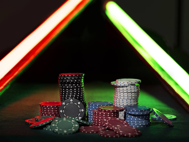 Close-up photo of a multicolored chips piles, some of them laying nearby on green cover of playing table, under green and red neon lights. Black background. Close-up. - Photo, Image