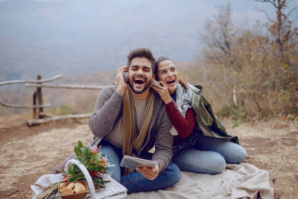 Beautiful smiling multicultural couple in love sitting on blanket at picnic in autumn, using tablet and listening music over headphones. Next to them are dog and basket with food. - Photo, Image