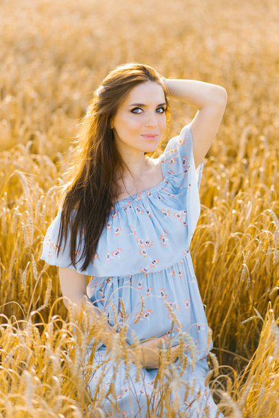 A pregnant woman in a blue dress holds her hand on her stomach in a wheat field at sunset - Foto, imagen
