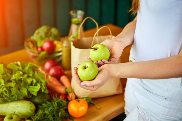 Young woman at kitchen taking out apple from grocery shopping paper bag with fruits and vegetables products.  - Foto, Bild