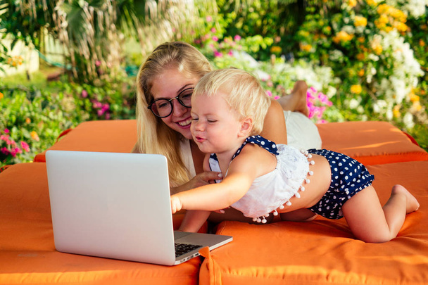 fair-haired one year old female kid looking at a laptop cartoon with mother blonde freelancer surfer on sunbeds resort summer beach.business woman freelancing and daughter relaxing by the sea - Фото, изображение