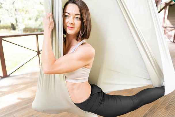 Pretty young woman practising anti-gravity fly yoga in white hammock. stretching exercises outdoors - Foto, Imagem
