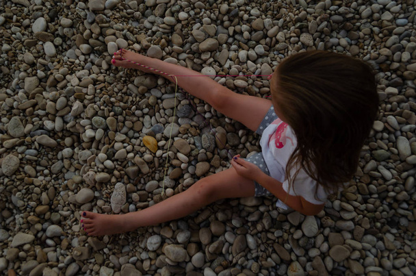 Vue du dessus d'une petite fille faisant bracelet en fil assis sur une plage de galets
 - Photo, image