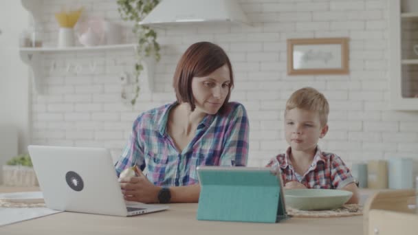 Attractive young mother and toddler son eating chocolate biscuits with their gadgets in bright kitchen - Filmagem, Vídeo