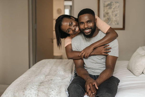 Portrait of a smiling young African American woman hugging her husband from behind while sitting in their pajamas on their bed in the morning - Фото, изображение